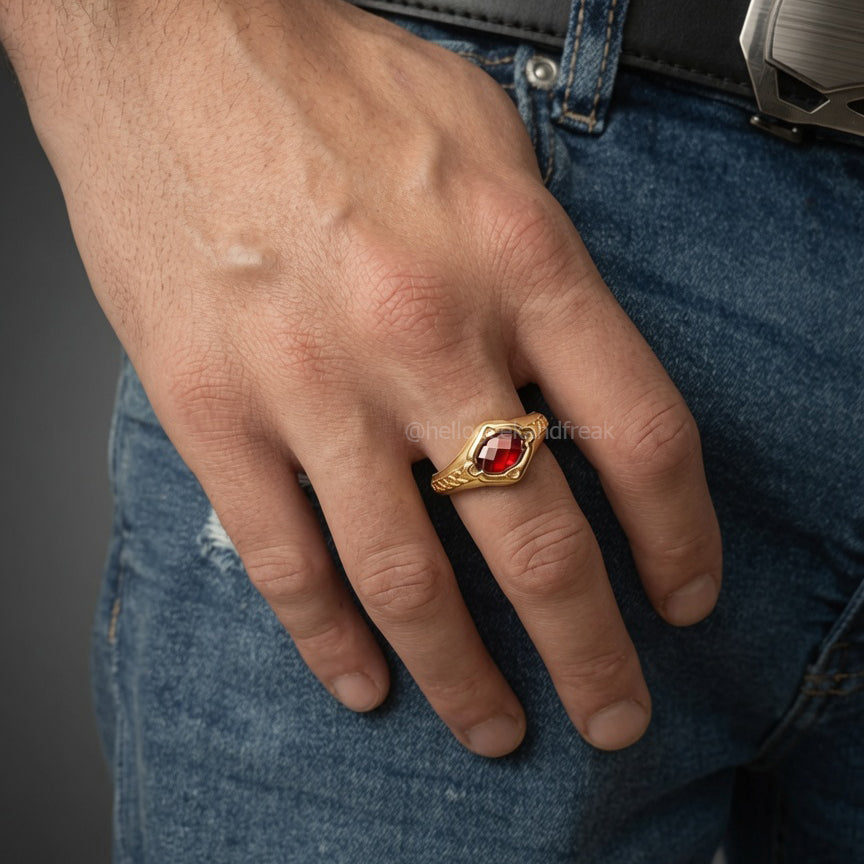 Anillo de metal dorado con piedra roja oscura y filigrana alrededor, en mano.